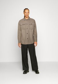 Bruine wollen blouse met knopen en twee borstzakken, gecombineerd met losse zwarte broek en zwarte schoenen, staand tegen een witte achtergrond.