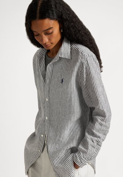 Γυναίκα με μακριά σγουρά μαλλιά, φοράει γκρι και λευκό πουκάμισο με κατακόρυφες ρίγες πάνω από γκρι τοπ, με τα χέρια στις τσέπες ανοιχτόχρωμου παντελονιού.
