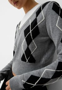 Grå stickad tröja med V-ringning, med svart och vitt argyle-mönster, ribbad nederkant och långa ärmar, mjuk textur.