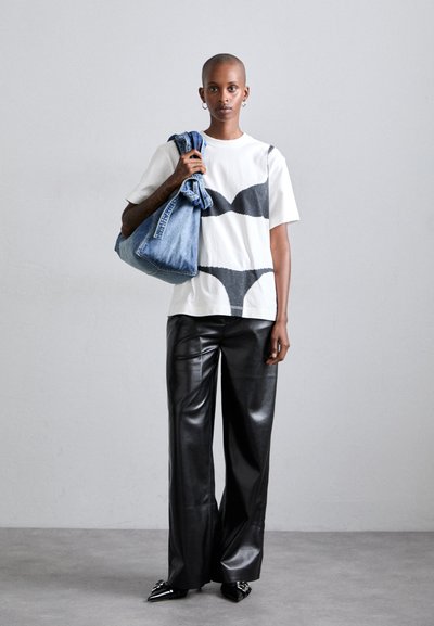 T-shirt bianco con motivo ondulato nero, pantaloni larghi in pelle nera e scarpe a punta nere, che tiene una borsa di jeans con jeans arrotolati.