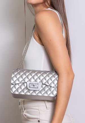 Donna che indossa una canotta bianca senza maniche e pantaloni beige tiene una piccola borsa trapuntata argento con tracolla a catena sulla spalla.