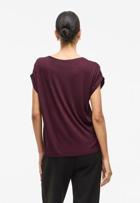 Burgunderfarget t-skjorte med korte ermer, avslappet passform, myk tekstur, rullet mansjett og vid halslinning, kombinert med svarte bukser.