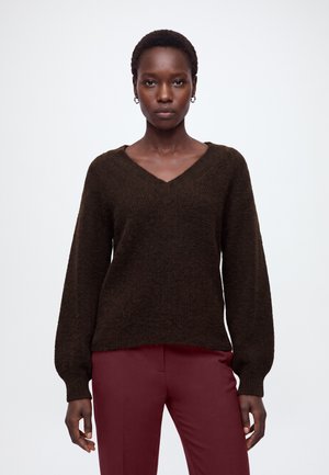 Femme aux cheveux courts portant un pull à col en V marron foncé et un pantalon grenat, debout devant un fond blanc uni.