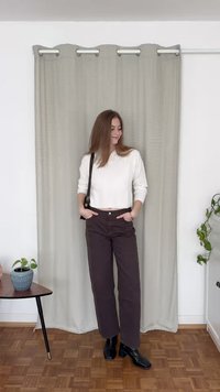 Camisola branca de manga longa canelada, calças largas castanho-escuras, botas de tornozelo pretas. De pé contra uma cortina cinza-clara com uma planta em vaso próxima.