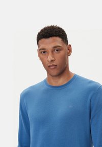 Maglione girocollo blu realizzato in tessuto liscio, con un piccolo logo ricamato sul petto. Maniche lunghe e dettaglio del collo a coste.