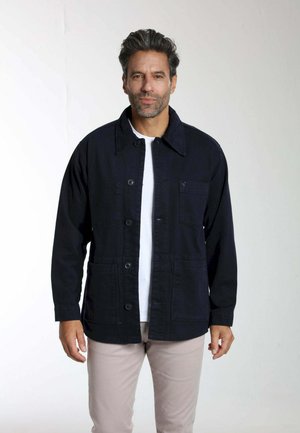 Homme aux cheveux noirs portant une veste noire boutonnée par-dessus une chemise blanche et un pantalon beige clair, debout devant un fond clair uni.