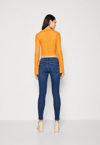Top corto de encaje naranja con mangas largas, combinado con jeans ajustados azul oscuro y zapatos de tacón bloque color beige claro. Diseño simple y ajustado.