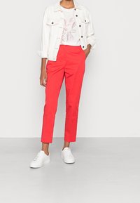 Pantalones rojos ajustados de algodón, combinados con una camisa blanca con un diseño de hojas, sobre la que se lleva una chaqueta de mezclilla clara y zapatillas blancas.