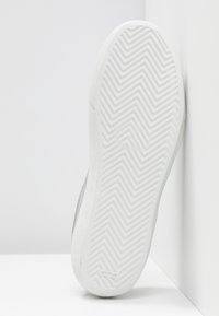 Semelle blanche de basket avec motif en chevrons, tenue à la verticale contre un mur et un sol blancs.