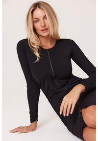 Czarna sukienka z długim rękawem, okrągłym dekoltem i zamkiem z przodu. Miękki materiał, dopasowany krój oraz drapowania w talii podkreślają sylwetkę.