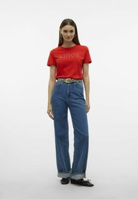 T-shirt en coton rouge avec impression "ADIOS", associé à un jean en denim bleu clair taille haute. Des ballerines noires et une ceinture à motif zèbre complètent le look.