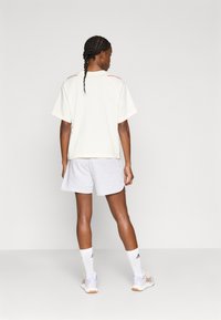 Ohlapna svetlo bež majica s kratkimi rokavi, sive kratke hlače, bele nogavice in športni čevlji; na ramenih so rdeči poudarki.