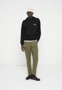 Νεαρός άνδρας που φοράει μπεζ σκούφο, μαύρο φούτερ με το κείμενο "Originals Studio", χακί παντελόνι και σκούρα καφέ μοκασίνια, στέκεται μπροστά σε έναν λευκό τοίχο.