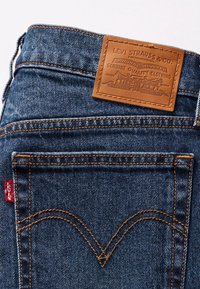 Vaqueros de mezclilla azul oscuro con un parche de cuero marrón con texto en relieve. Costuras en naranja contrastante y una etiqueta roja de Levi's en el bolsillo trasero.
