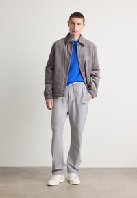 Chaqueta de pana gris con cuello, suéter azul debajo, pantalones de chándal gris claro y zapatillas blancas. Diseño minimalista con detalles de logo.