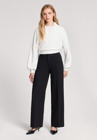 Biały sweter prążkowany z dekoltem w serek, w połączeniu z czarnymi spodniami o szerokich nogawkach i wysokim stanem. Modelka nosi czarne szpilki.