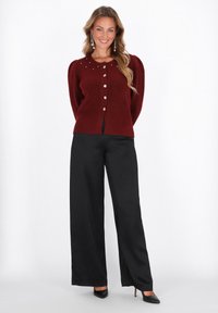 Donna sorridente che indossa un maglione bordeaux con bottoni e dettagli in perle, pantaloni neri a gamba larga, tacchi neri e orecchini lunghi, su sfondo bianco.