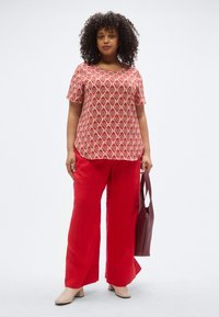 Κορυφή με κόκκινο μοτίβο και κοντά μανίκια, συνδυασμένη με φαρδιά κόκκινη παντελόνα. Αξεσουάρ περιλαμβάνουν μια μπορντό τσάντα και μπεζ παπούτσια με τακούνι τύπου block.