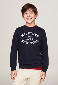 Felpa navy con la scritta bianca "HILFIGER EST. 1985 NEW YORK", polsini a costine e colletto girocollo. Indossata sopra una camicia rossa, abbinata a pantaloni chiari.