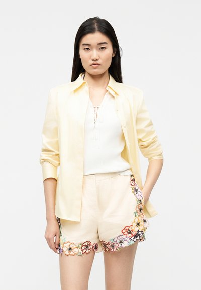 Jeune femme portant une chemise jaune pâle sur un haut blanc et un short beige avec une broderie florale colorée le long des bords.