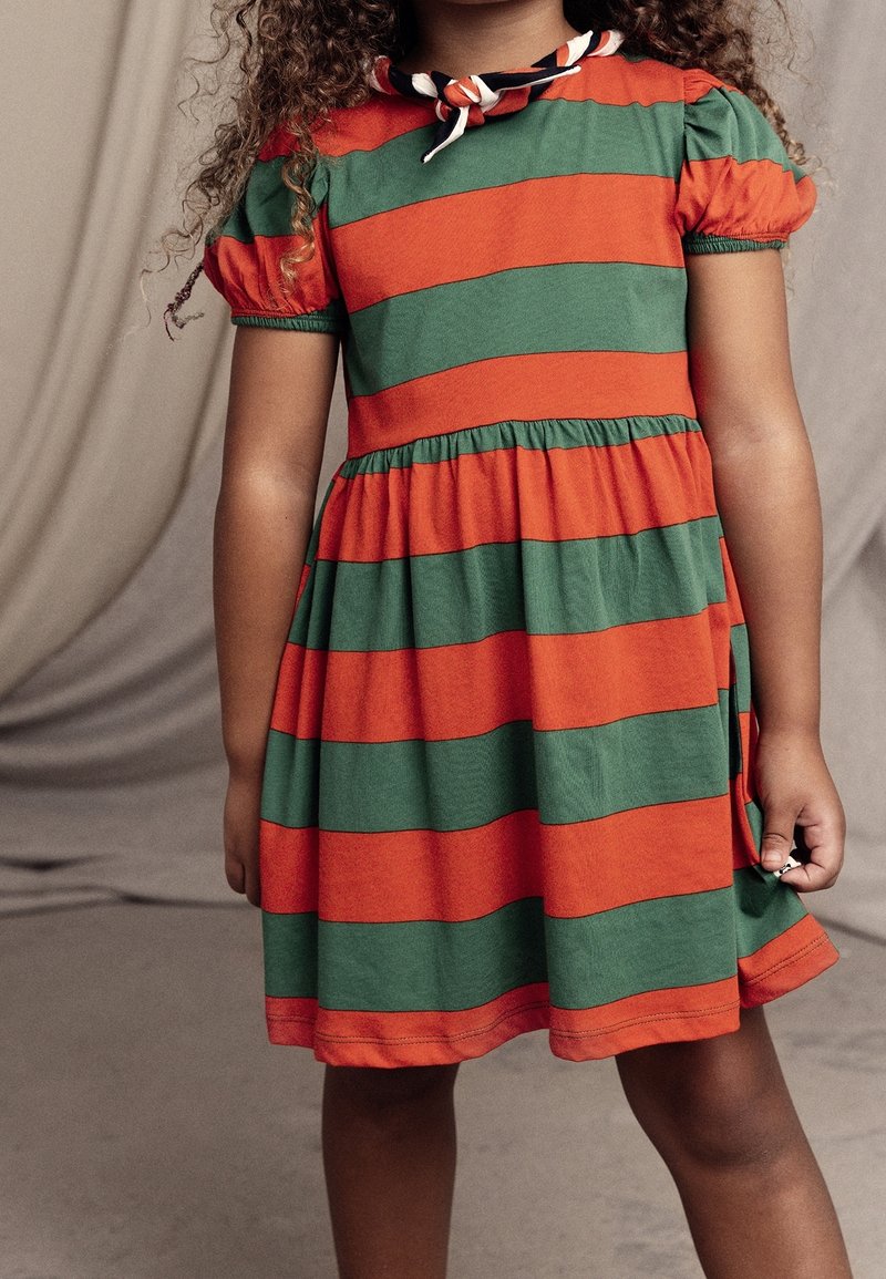 Enfant portant une robe à larges rayures orange et verte avec des manches bouffantes et un foulard noué autour du cou.