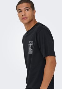 T-shirt preta de algodão com um gráfico branco impresso do Homem Vitruviano e texto na frente. Mangas curtas, gola redonda, corte descontraído.