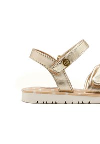 Sandal in metallo oro con due cinturini, suola piatta bianca e soletta decorata con stelle. L'hardware include un logo metallico rotondo.