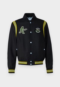 Veste universitaire noire avec des boutons-pression bleus, des accents rayés jaunes et bleus, et des patchs brodés d'un joueur de tennis vert et d'un logo.