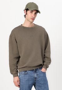 Sweatshirt vert olive avec un col rond et des épaules tombantes, accompagné d'une casquette assortie. Un jean bleu clair complète la tenue.