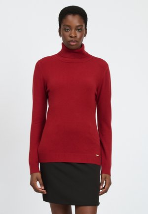 Maglione a collo alto rosso in morbido tessuto a maglia con orlo a coste. Presenta maniche lunghe e un piccolo logo dorato sul lato inferiore.