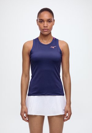 Une femme se tient face à l’avant, vêtue d’un haut de sport sans manches bleu marine et d’une jupe de tennis blanche, sur un fond clair uni.