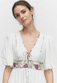 Weißes Kleid mit Puffärmeln, einem V-Ausschnitt und einem taillierten Blumenmuster. Der Stoff hat eine leicht strukturierte Oberfläche.