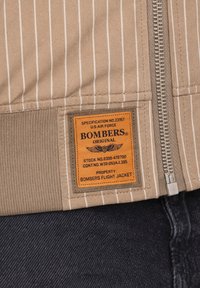 Veste rayée beige avec une fermeture éclair métallique et un patch orange sur lequel est inscrit "Bombers Original" ainsi que les spécifications de l'US Air Force.