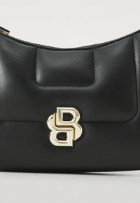Sac à main en cuir noir avec une texture lisse, une forme arrondie et un accent logo en ton doré. Fermeture éclair avec des détails de couture visibles.