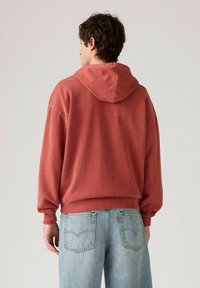Felpa rossa con cappuccio, dal fit rilassato e polsini a coste, abbinata a jeans azzurri chiari con dettagli di cucitura sulle tasche posteriori.