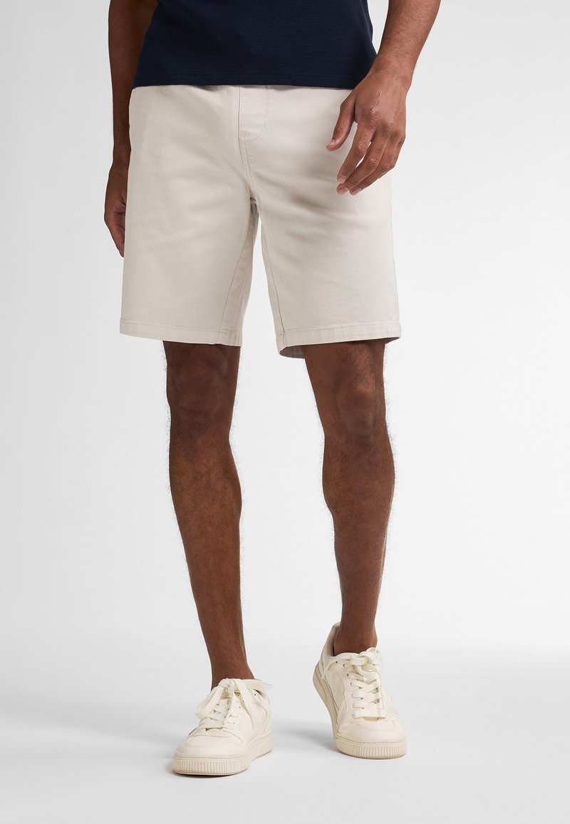 Helle beigefarbene Baumwollshorts mit geradem Schnitt, seitlichen Taschen und einem sauberen Saum, kombiniert mit weißen Sneakers, vor einem neutralen Hintergrund.