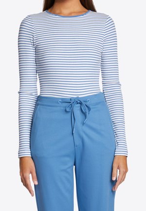 Femme portant un haut ajusté à manches longues à rayures horizontales blanches et bleues, rentré dans un pantalon bleu taille haute avec une ceinture à cordon nouée.