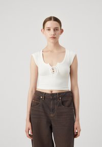 Hvit ribbet crop top med scoop neckline og snøring foran, kombinert med mørkebrune høytlivsjeans med oransje søm.
