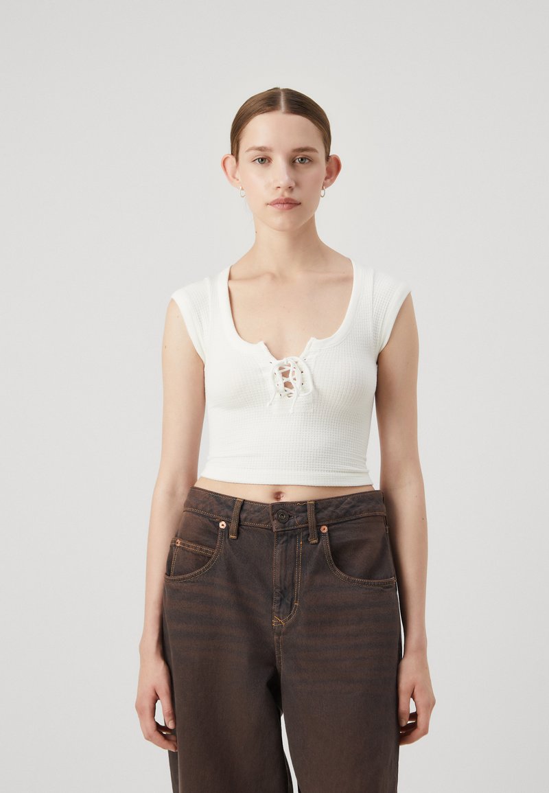 Hvit ribbet crop top med scoop neckline og snøring foran, kombinert med mørkebrune høytlivsjeans med oransje søm.