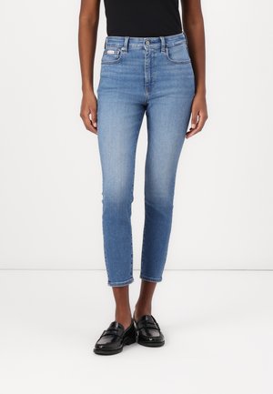 Dżinsy slim fit w jasnoniebieskim kolorze, noszone z czarnymi loafersami i czarnym topem, pokazane od talii do stóp na osobie stojącej na białym tle.