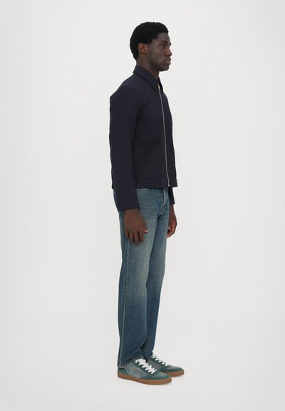 Giacca zip con collo di colore blu navy, due tasche frontali, indossata con jeans in denim azzurri e scarpe da ginnastica verdi con lacci bianchi e suole marroni.