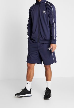 Homme portant une veste de sport bleu marine et un short assorti à rayures blanches, des baskets noir et blanc, et des chaussettes blanches Puma, debout.