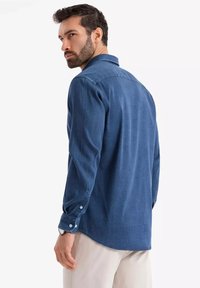 Camicia in denim blu con maniche lunghe, colletto con bottoni e polsini con bottoni. Presenta una texture liscia e un orlo leggermente curvato.