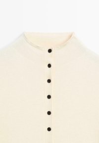 Pull col roulé de couleur crème avec une texture côtelée et un ourlet droit. Doté d'une patte de boutonnage à cinq boutons noirs au centre.