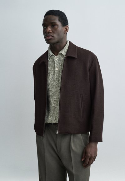 Jeune homme portant une veste zippée marron foncé par-dessus une chemise grise texturée boutonnée et un pantalon gris plissé, debout devant un fond uni.
