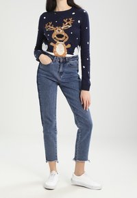 Sweter dziergany w kolorze granatowym z wzorem renifera, jasnokremowe akcenty, białe kropki; zestawiony z wysokim stanem, skróconymi niebieskimi dżinsami i białymi tenisówkami.