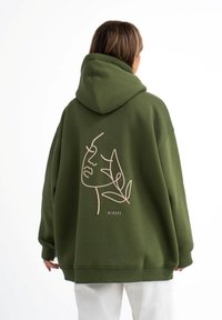 Groene oversized hoodie met een capuchon, met een minimalistische lijntekening van een gezicht en bladeren in beige op de achterkant. Gemaakt van een katoenmix.