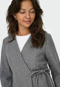 Grijze jasje in wikkelstijl gemaakt van zachte viltstof. Heeft een strikceintuur in de taille en lange mouwen, gelaagd over een wit shirt.