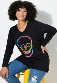 Schwarzes Langarm-T-Shirt mit einem bunten Totenkopf-Graphic; kombiniert mit blauen Jeans mit Farbspritzern; Modell sitzt auf einem gelben Hocker.