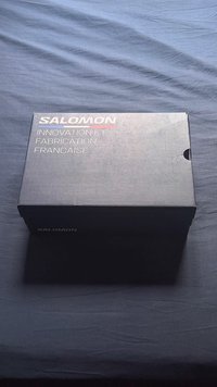 Caixa de sapatos retangular preta com um acabamento mate. Logótipo "SALOMON" em prateado, com texto em francês "INNOVATION ET FABRICATION" impresso em branco.